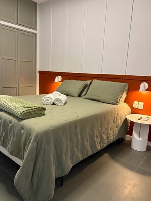 A neatly made bed with a green bedspread, two pillows, and a folded blanket. There is a bedside table with a lamp and a small clock. The room has a wooden headboard and a white wall with a door. - Guarapari Center Aparts (Guarapari)