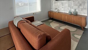 Egy nappali, amelyben van egy kanapé, a falra szerelt televízió, a fal pedig kőmintázatú, emellett mennyezeti ventilátor és egy nagy ablak is található benne.