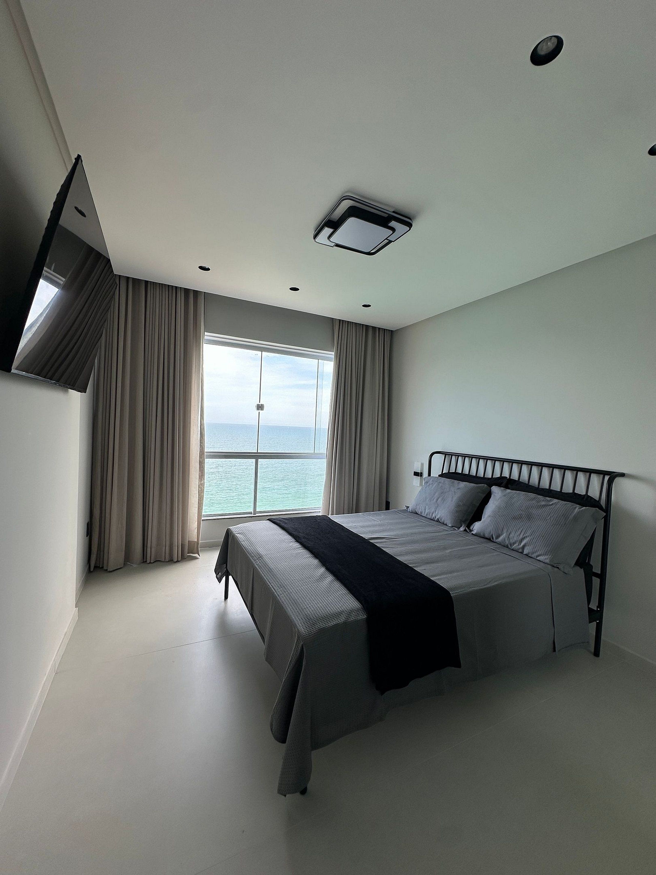 A bedroom with a bed, a window with curtains, a flat-screen TV mounted on the wall, and a ceiling light fixture.