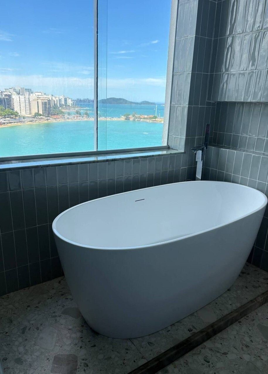 Ein modernes Badezimmer mit einer großen Badewanne, Blick auf Strand und Stadtlandschaft durch ein großes Fenster, und dunkel geflieste Wände.