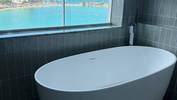 Ein modernes Badezimmer mit einer großen Badewanne, Blick auf Strand und Stadtlandschaft durch ein großes Fenster, und dunkel geflieste Wände.