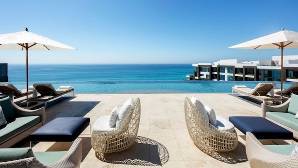Un espace aménagé au bord de la piscine, avec des fauteuils de relaxation en osier et des parasols, offrant une vue sur un océan d’un bleu limpide et un bâtiment moderne doté de balcons.