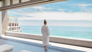 Une personne vêtue d’une longue robe blanche se tient sur un balcon surplombant la mer. On aperçoit également un bâtiment moderne et des chaises longues.