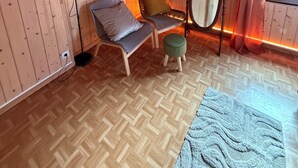 Et værelse med trævægge og -loft, et spejl, to stole med hynder, en gulvlampe og et vindue med gardiner.