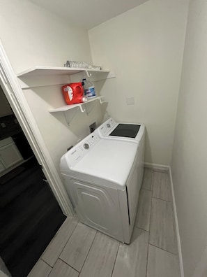 A compact laundry room with a front-loading washing machine and a shelf above containing cleaning supplies. - Econo Comfort near TCU & Downtown (Fort Worth)