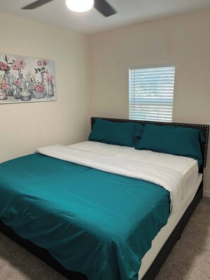A bedroom with a bed, teal bedding, a ceiling fan, a window with blinds, and a floral painting on the wall. - Comfortable & Modern Home near TCU-Downtown-Shops (Fort Worth)