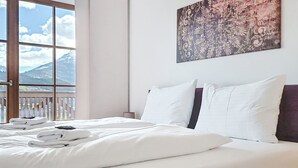 Un lit bien fait, avec du linge de lit blanc et des oreillers, une tête de lit en bois et une grande fenêtre donnant sur les montagnes.
