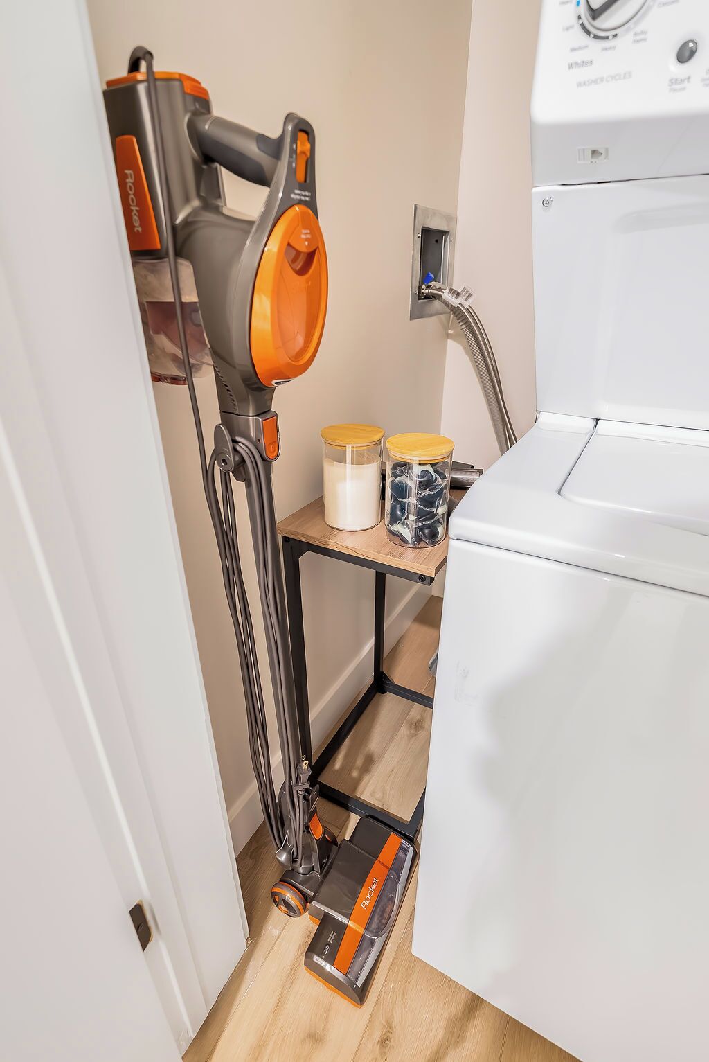 A compact laundry area with a washing machine, a vacuum cleaner, and storage jars on a small table.