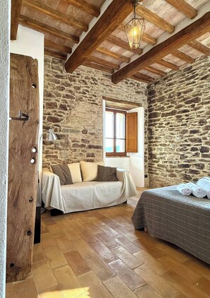 A rustic room with exposed stone walls, a wooden ceiling, a bed, a sofa, and a window with wooden shutters. - Torre Apartment - Spacious medieval living at Torre della Soara (Umbria)