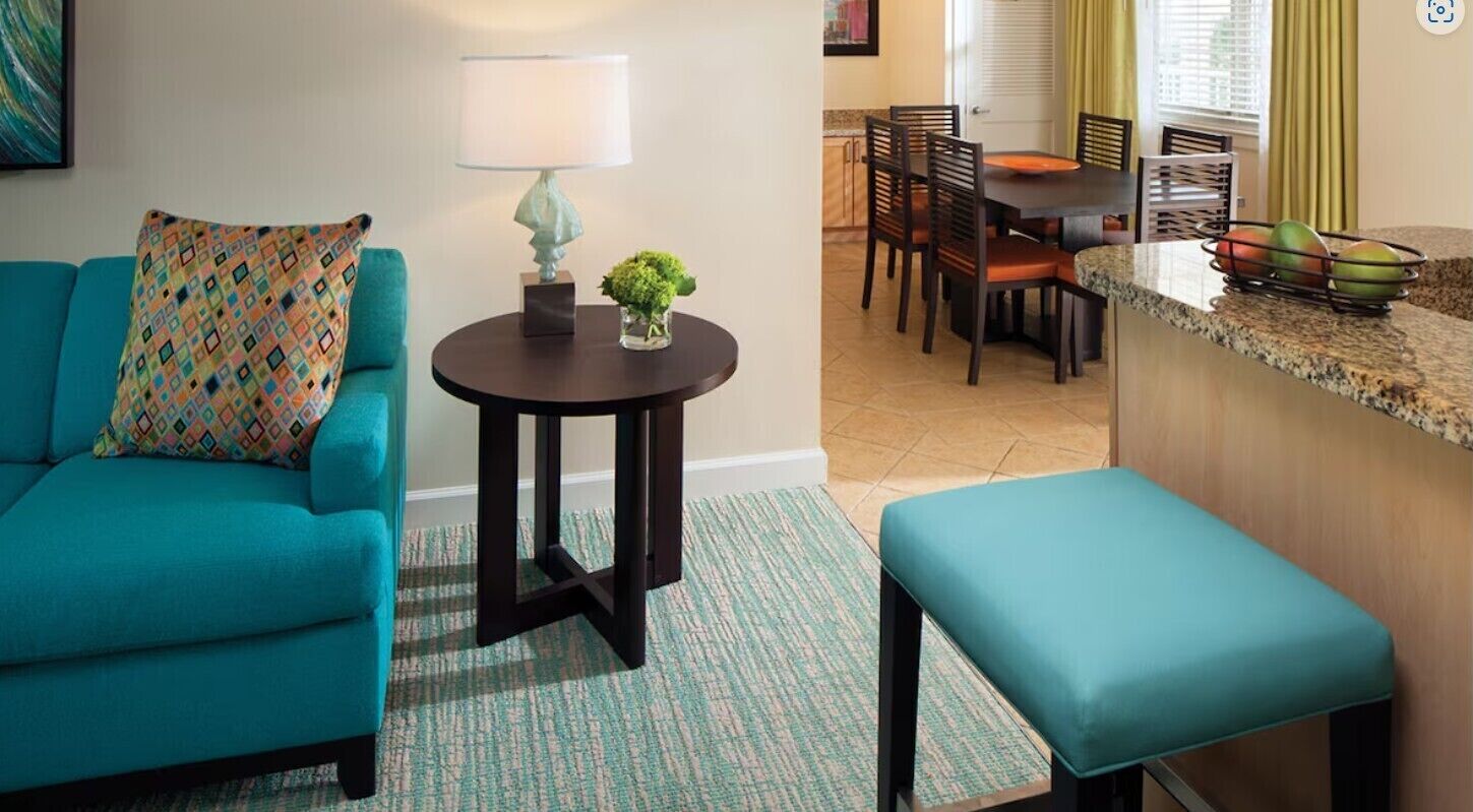 A living room with a teal sofa, a small round table with a lamp, a bench, and a kitchen area with a counter and chairs.