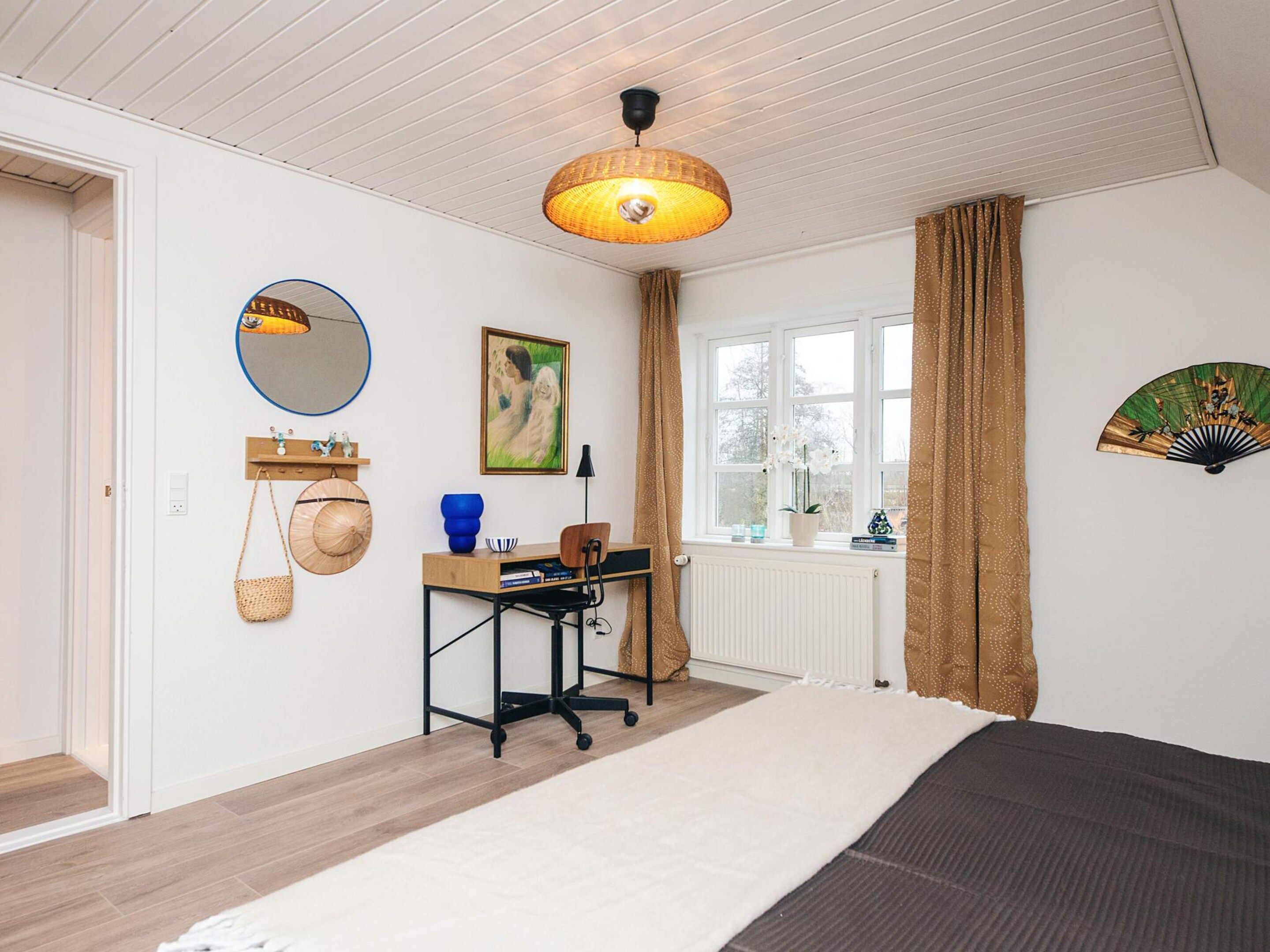 A bedroom with a bed, a desk with a chair, a window with curtains, a wall-mounted fan, and a mirror.