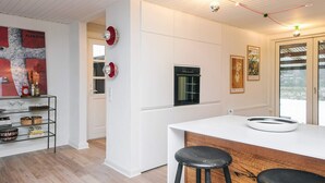 Et moderne køkken med hvide skabe, en sort ovn og en hvid bordplade. To sorte barstole og en hvid hylde med forskellige ting.
