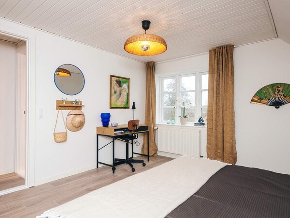 Une chambre à coucher avec un lit, un bureau avec une chaise, une fenêtre avec des rideaux, un ventilateur fixé au mur, un miroir et un tableau.