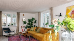 En moderne stue med en gul sofa, en sort lænestol, en rød måtte og store vinduer med gardiner.