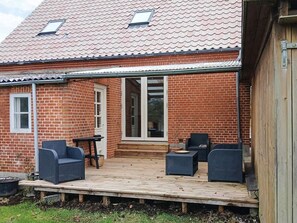 Een bakstenen huis met een houten terras, twee rieten stoelen, een tafel en een schuifdeur met glas.