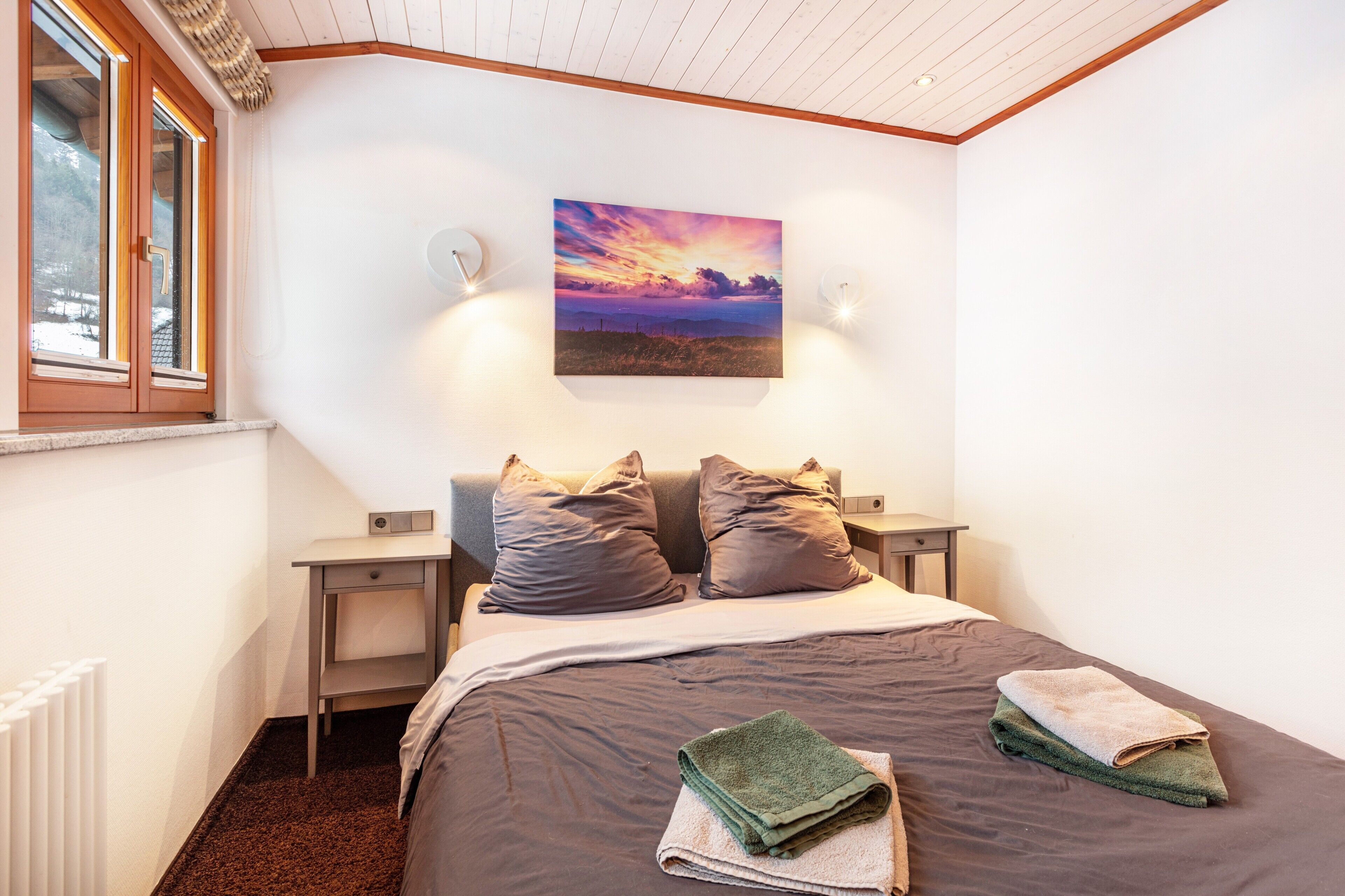 A bedroom with a bed, two bedside tables, a nightstand, a window, and a painting on the wall.
