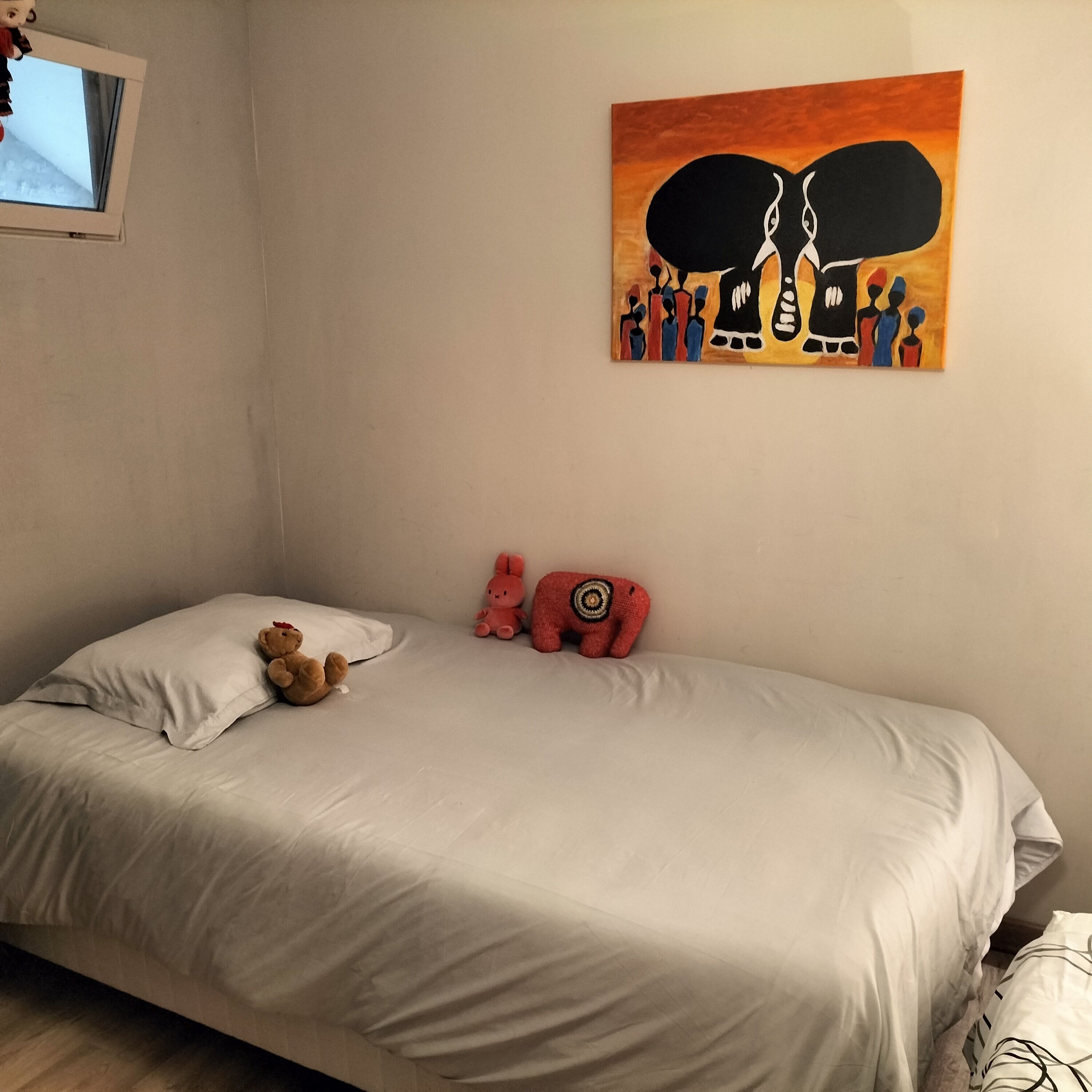 Un dormitorio con una cama, almohadas, un oso de peluche, un conejo rosa y un elefante rojo. Hay un cuadro en la pared.