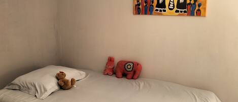 Un dormitorio con una cama, almohadas, un oso de peluche, un conejo rosa y un elefante rojo. Hay un cuadro en la pared.