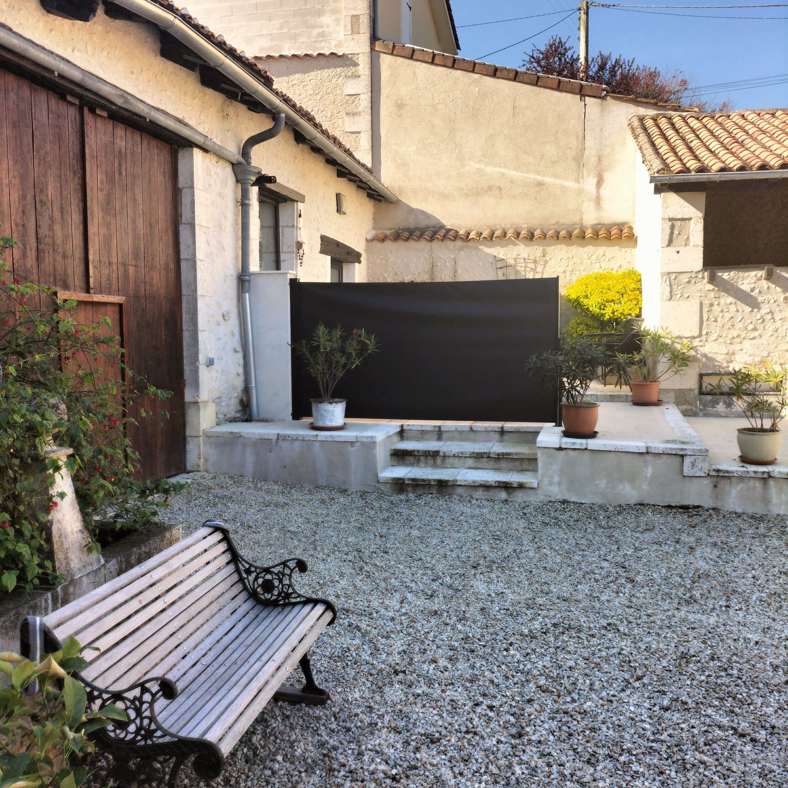 Un patio de grava con un banco de madera, una pantalla negra y plantas en macetas.