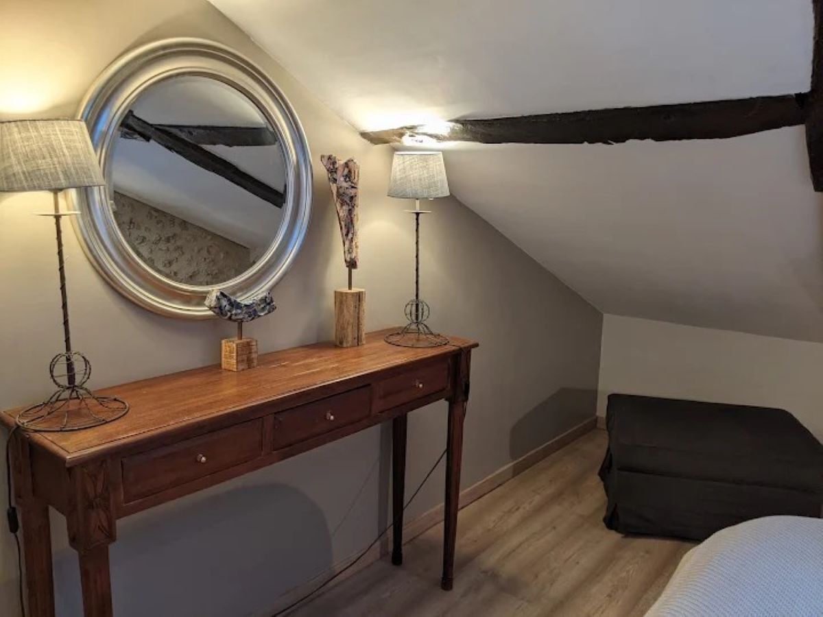 A cozy attic bedroom with a wooden dresser, a round mirror, two lamps, and a black sofa.