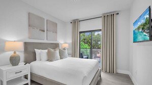 A modern bedroom with a large bed, a nightstand, a lamp, and a view of greenery through the window. - Miami Palm Villas - Family - Entire 8BR-6BA (Miami)