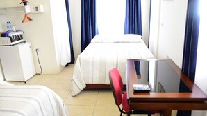 Habitación de hotel con una cama, un escritorio con una silla, un refrigerador pequeño y una repisa con una botella de agua y una radio.