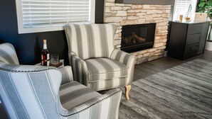 A living room with a stone fireplace, striped armchairs, a dark wooden cabinet, and a window with blinds. - New Luxury 3-bedroom Cabin in Big Bear Lake with Hot tub,EV Charger,Pool Table (Big Bear Lake)