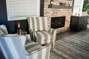 A living room with a stone fireplace, striped armchairs, a dark wooden cabinet, and a window with blinds. - New Luxury 3-bedroom Cabin in Big Bear Lake with Hot tub,EV Charger,Pool Table (Big Bear Lake)