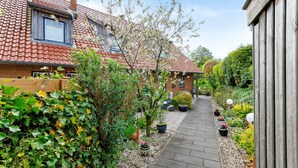A narrow pathway flanked by houses with red-tiled roofs and a garden with potted plants.