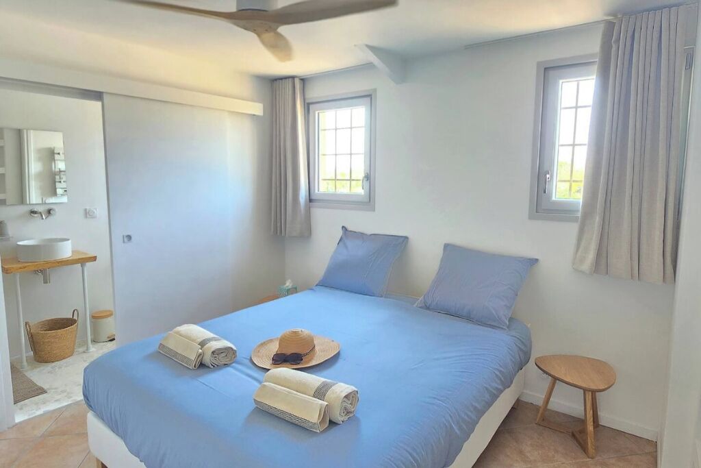 A bedroom with a bed, two windows, a ceiling fan, a bedside table, and a small stool.