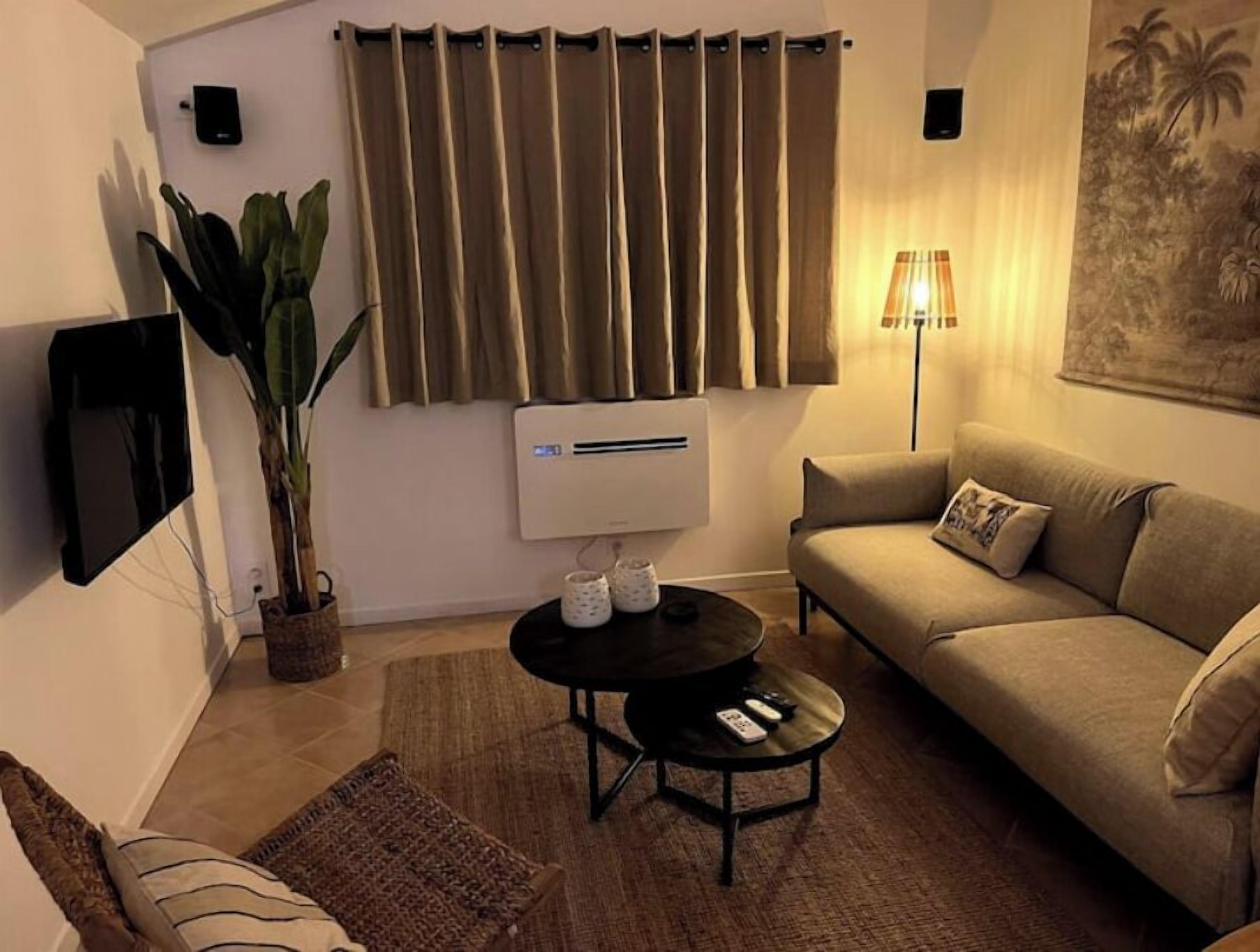 A living room with a beige sofa, a coffee table, a TV mounted on the wall, an air conditioning unit, and a plant.