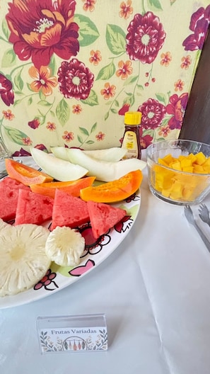 A plate of assorted fruits including watermelon, pineapple, and mango, with a bottle of honey and a glass of orange juice on a table with a floral tablecloth. - Caraiva dos Sonhos (Porto Seguro)