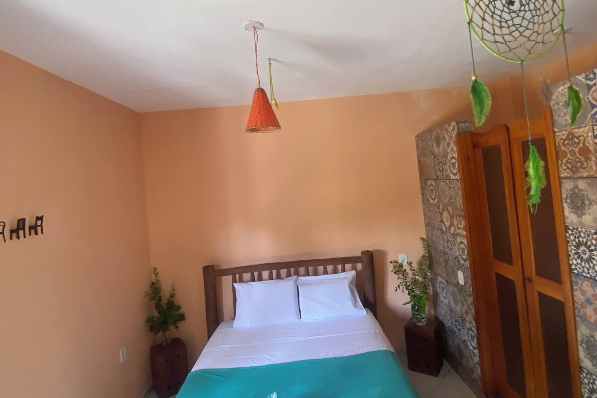 Un lit bien fait, avec une tête de lit en bois, une couvre-lit verte et un oreiller blanc. Une lampe orange est suspendue au plafond et un attrape-rêves vert est accroché au mur.