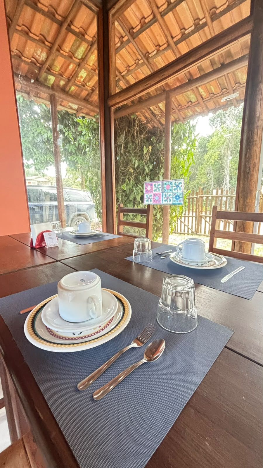 Mesa de comedor servida con una taza blanca, un platillo y cristalería, con vista a la vegetación exterior.