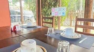 Mesa de comedor servida con una taza blanca, un platillo y cristalería, con vista a la vegetación exterior.