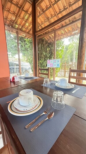 Mesa de comedor servida con una taza blanca, un platillo y cristalería, con vista a la vegetación exterior.