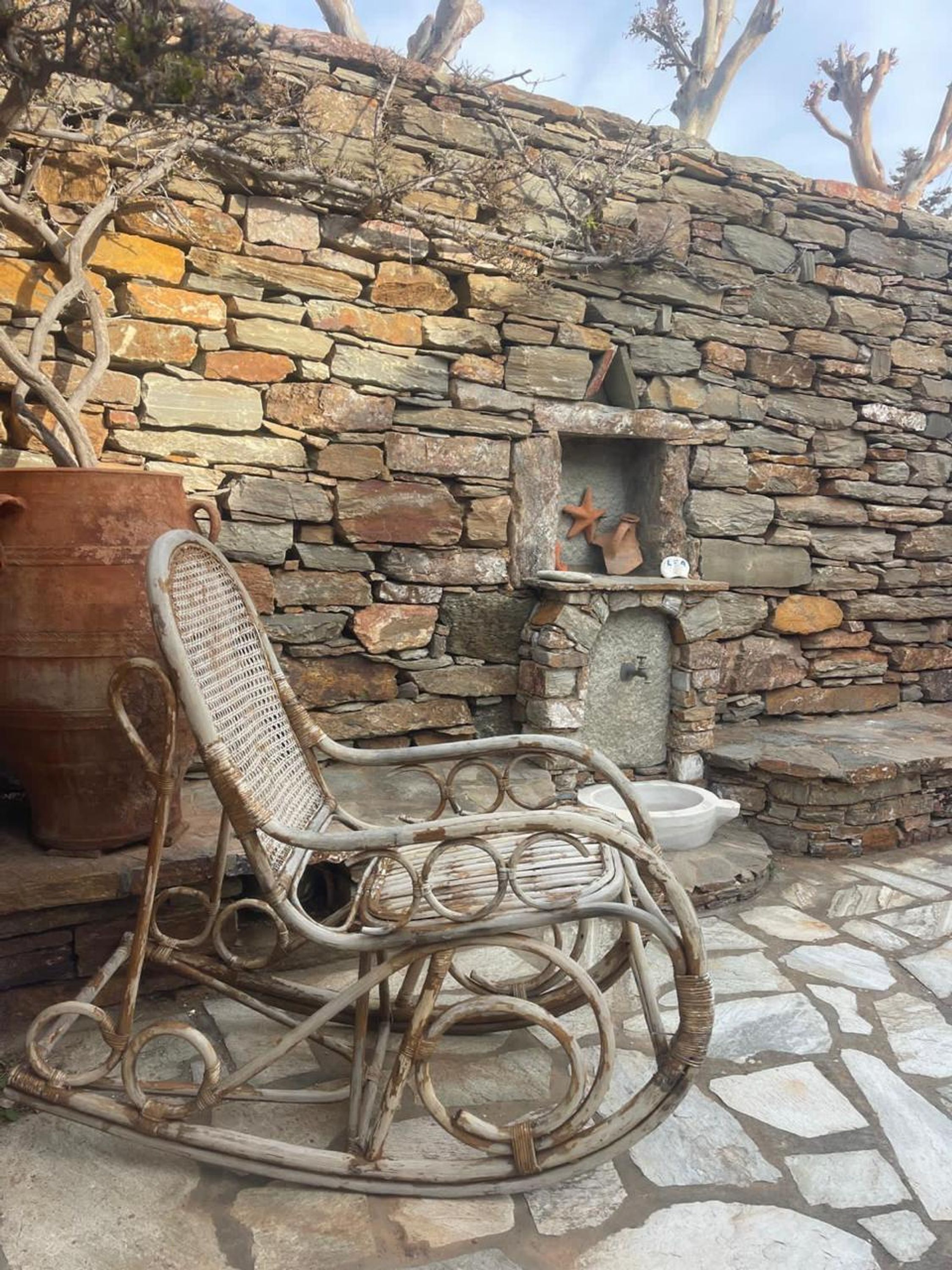 Un'area salotto esterna rustica con un muro in pietra, una sedia in vimini, una pentola di metallo e un piccolo ripiano con oggetti decorativi.