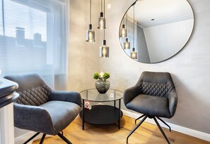 Ein modernes Zimmer mit zwei grauen Polsterstühlen, einem Glastisch mit einer Vase, einem runden Spiegel und Pendelleuchten.