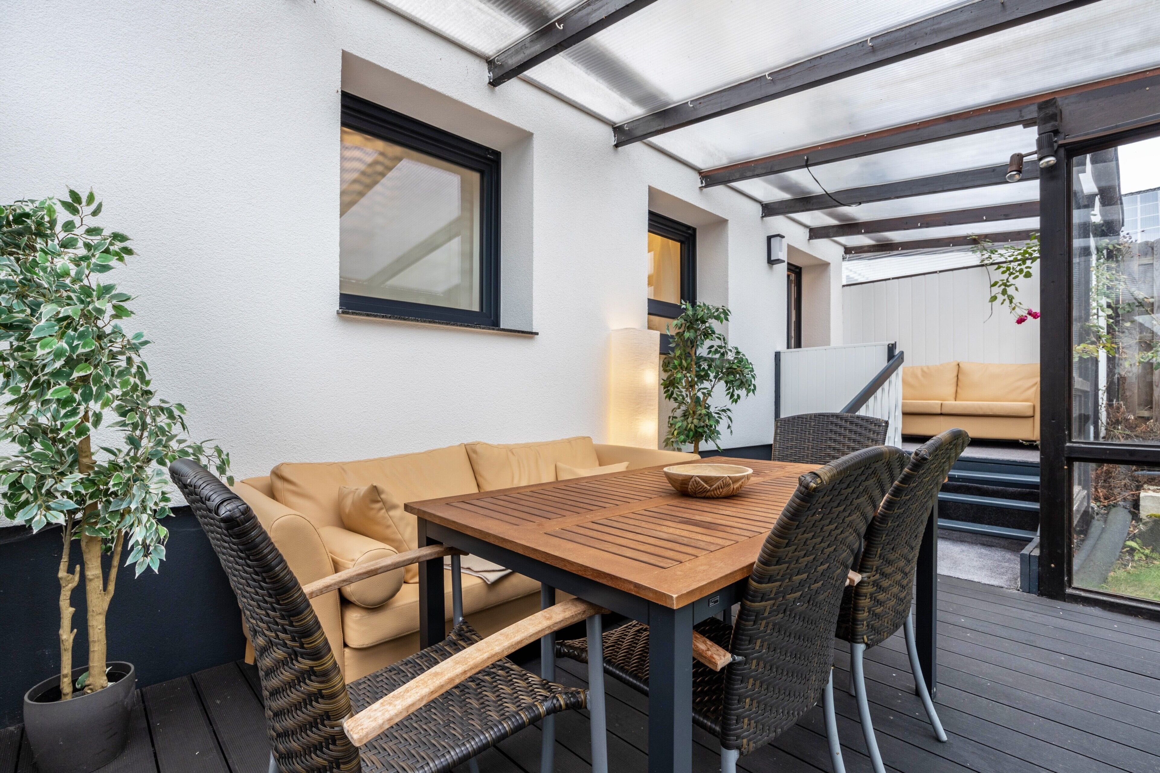 An outdoor patio with a wooden table, wicker chairs, a beige sofa, and potted plants.
