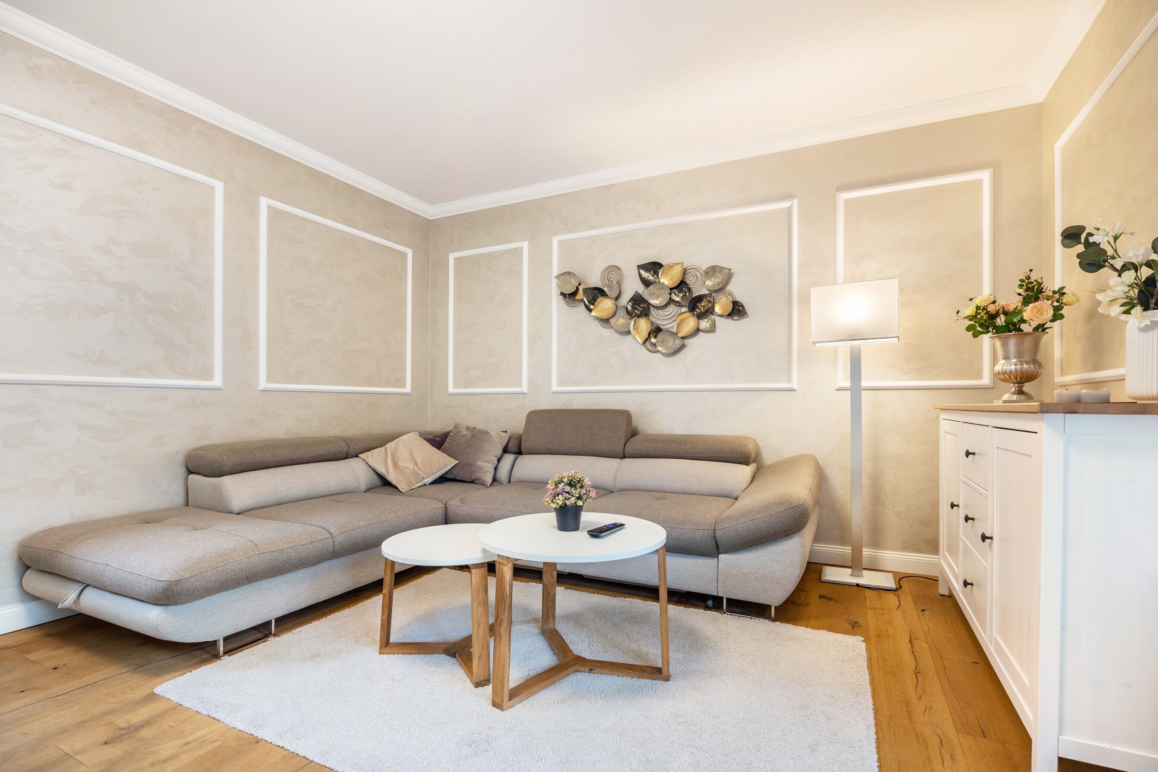 Ein modernes Wohnzimmer mit einem Ecksofa, zwei runden Tischen, einer Wanddekoration, einer Stehlampe und einem Schrank.