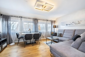 Ein modernes Wohnzimmer mit einem grauen Sofa, einem Esstisch mit Stühlen und einem großen Fenster mit Vorhängen.