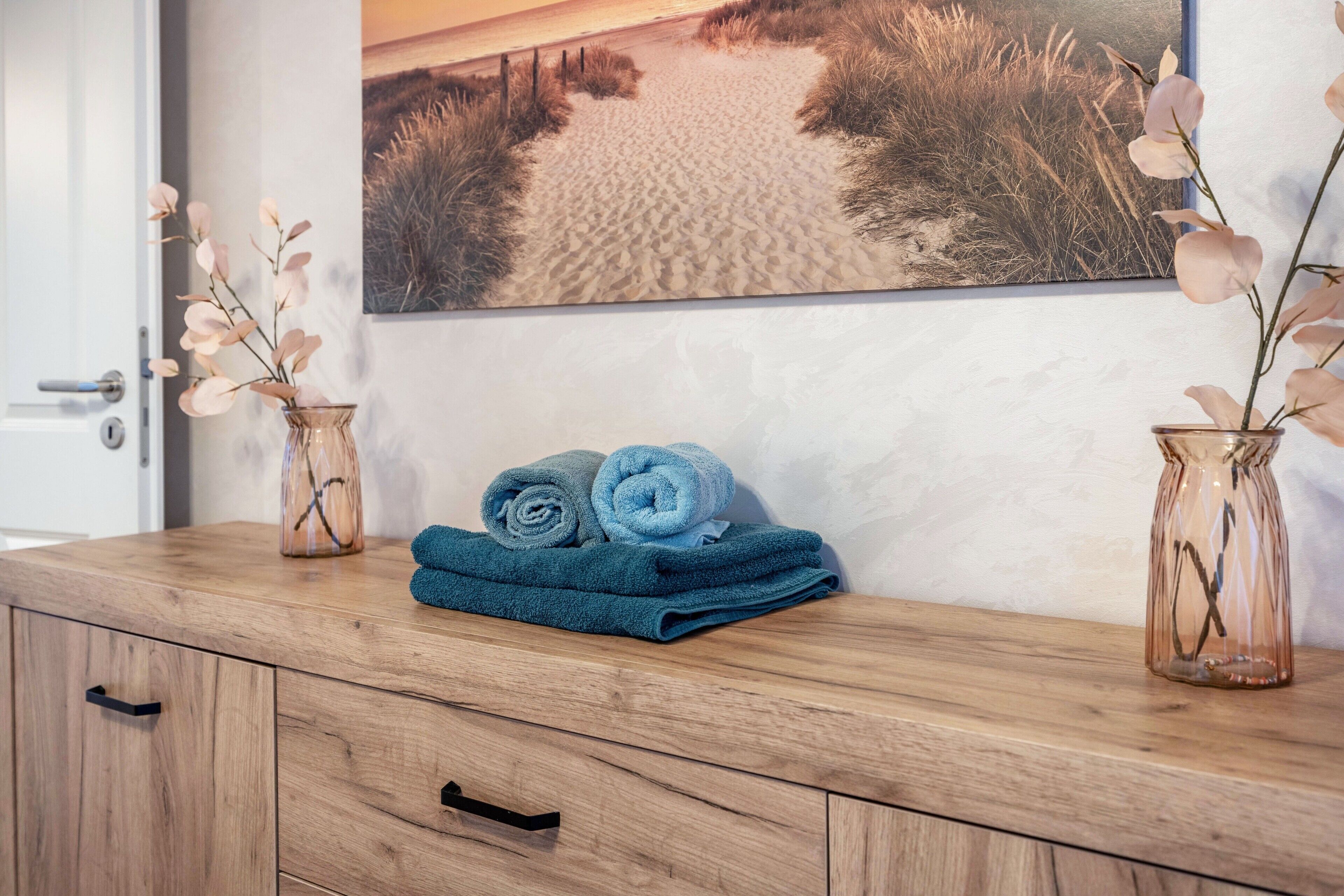 A wooden vanity with a rolled blue towel, two glass vases with dried flowers, and a beach-themed painting on the wall.