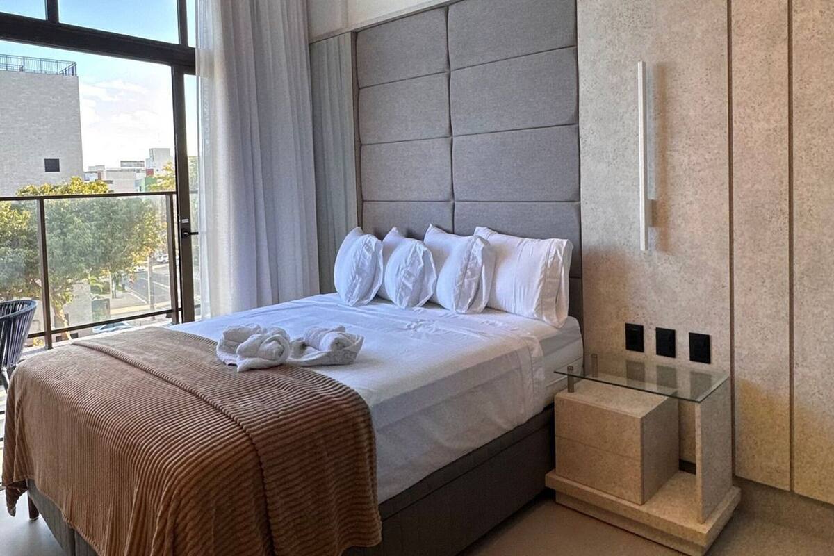 A modern bedroom with a large bed, a glass bedside table, a built-in wardrobe, and a view through a large window.