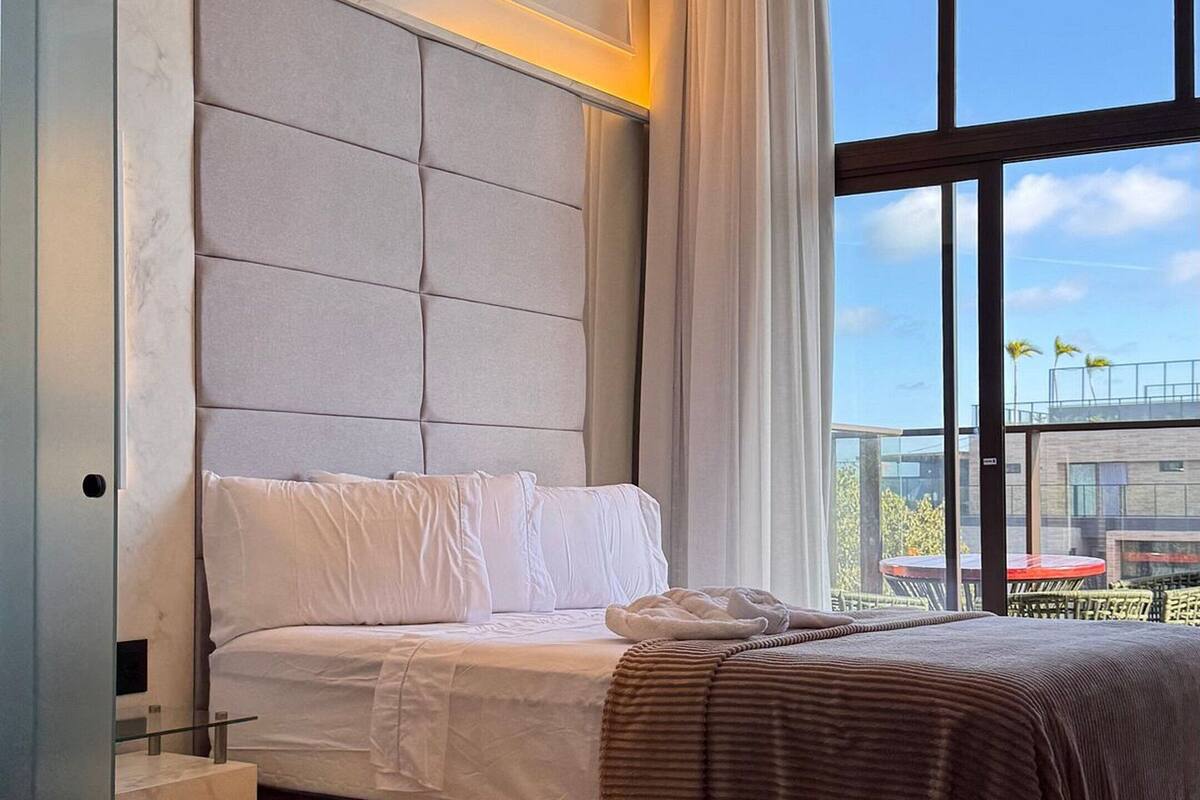 A modern bedroom with a large bed, a nightstand, a bedside table, and a view of the outdoors through large windows.