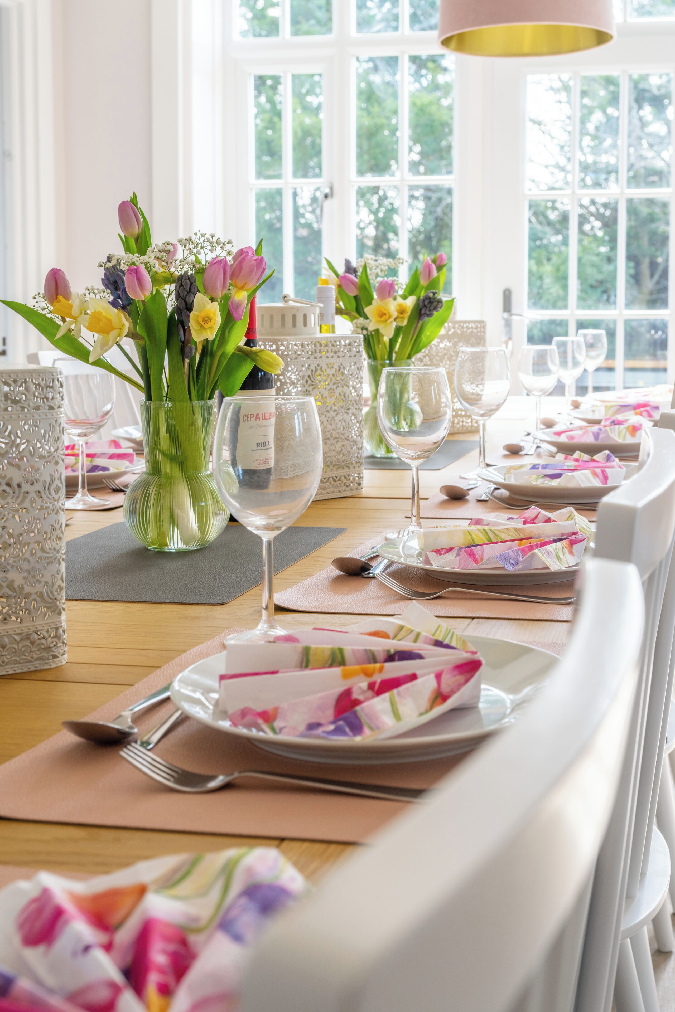 Une table à manger dressée avec des assiettes, des couverts et des verres, agrémentée de vases de fleurs et d’une bouteille de vin.