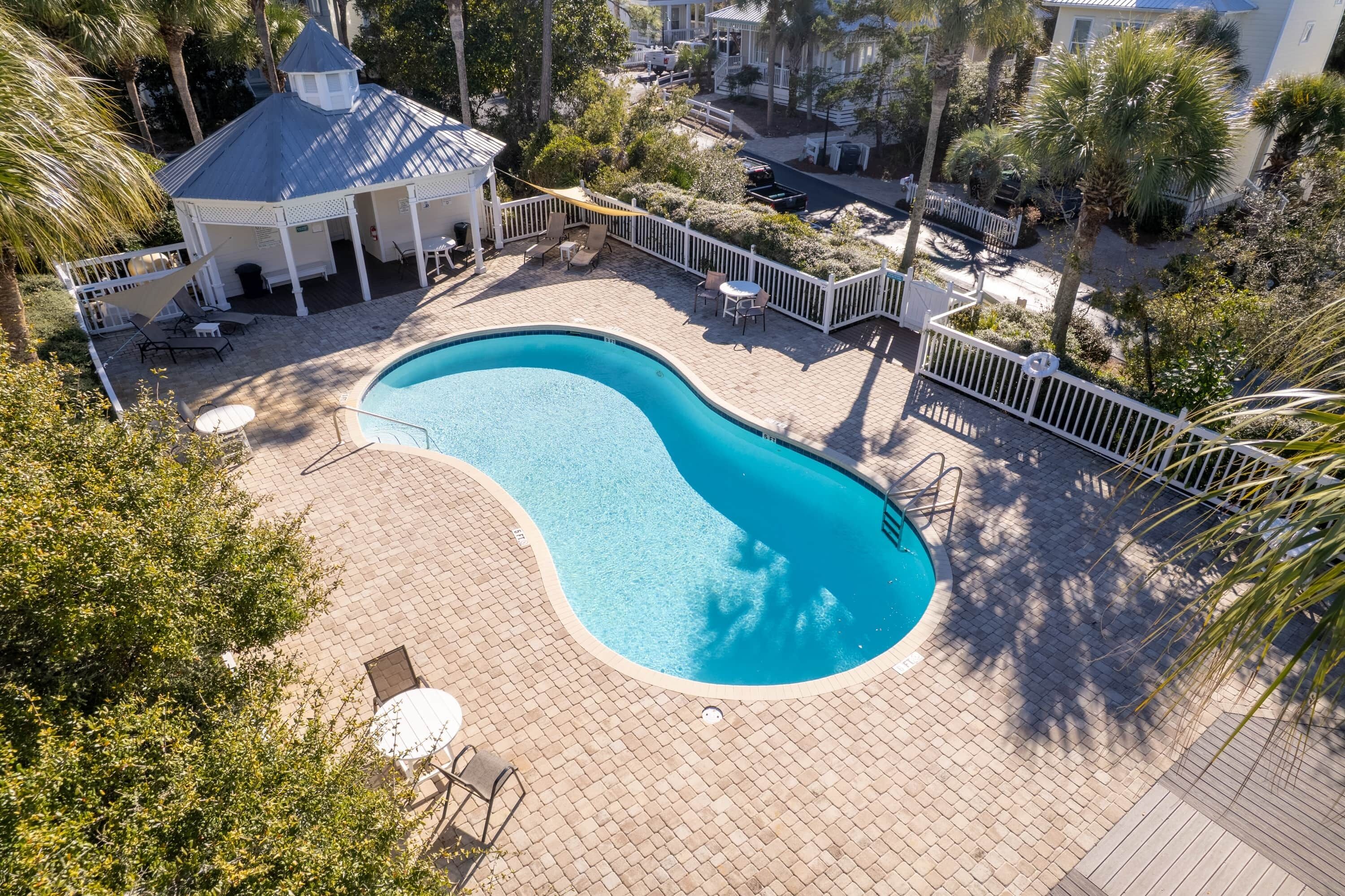 A swimming pool with a curved edge, surrounded by a white fence and a gazebo.
