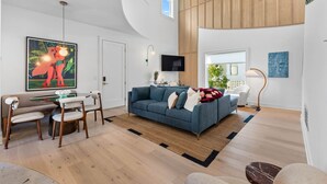 Uma sala de estar moderna com um sofá azul, piso de madeira, um espaço de jantar com mesa de vidro e um quadro grande.