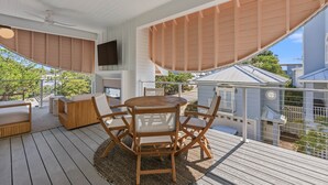 Um deck coberto ao ar livre com uma mesa redonda de madeira e cadeiras, um ventilador de teto e uma televisão fixada na parede.