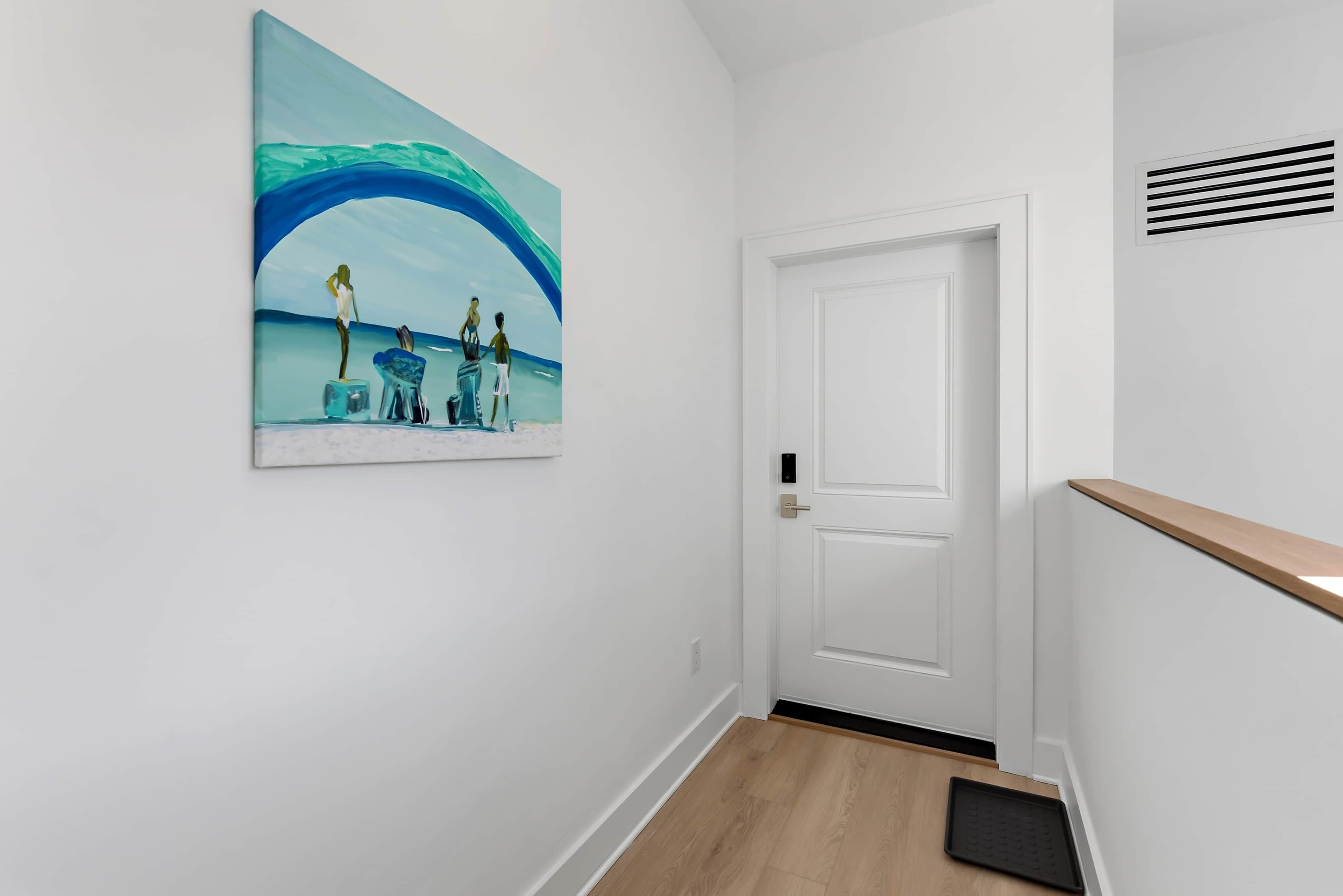 A hallway with a white door, a wooden floor, and a painting of a beach scene.