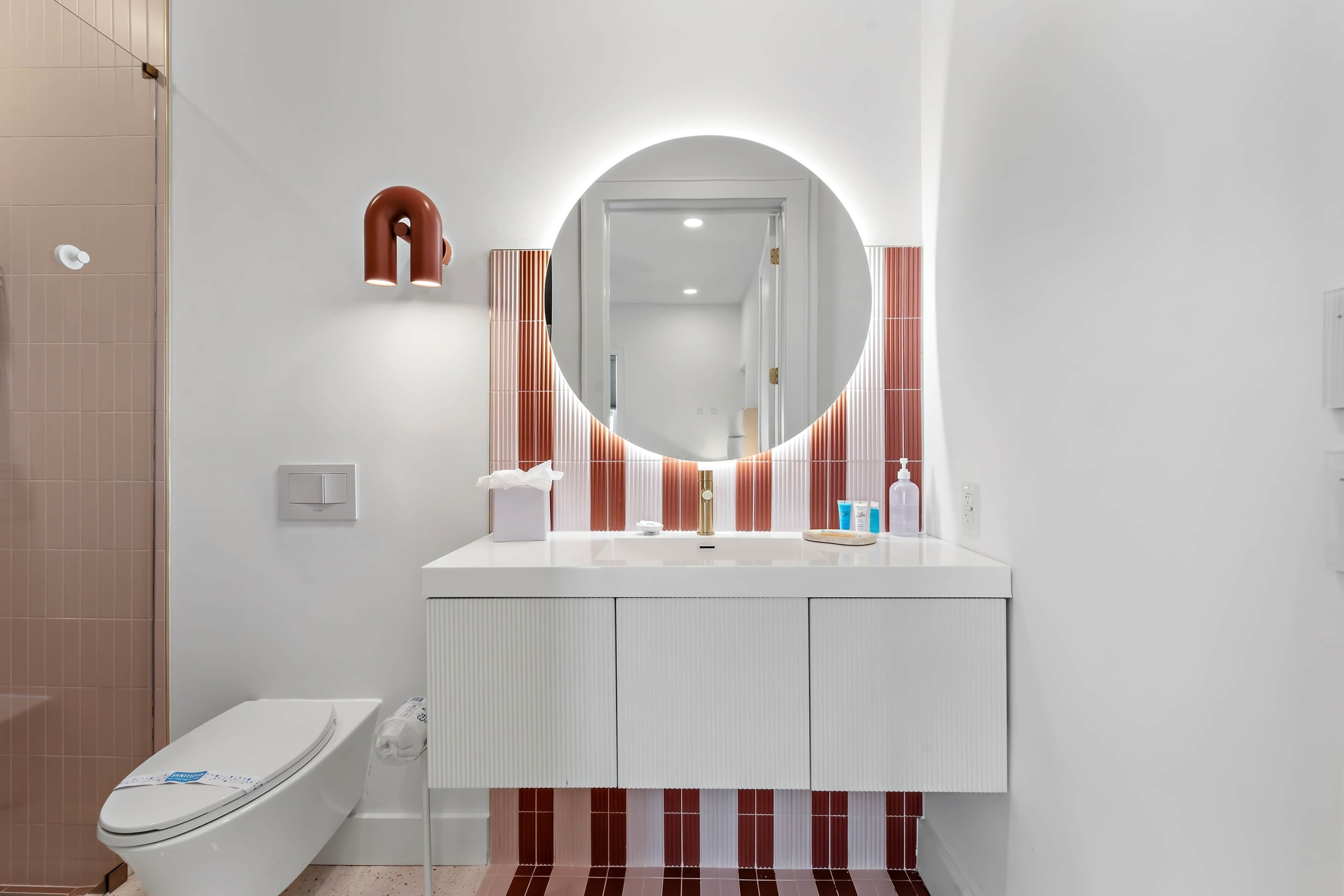 A modern bathroom with a white vanity, a round mirror with a light, striped wall tiles, and a toilet.